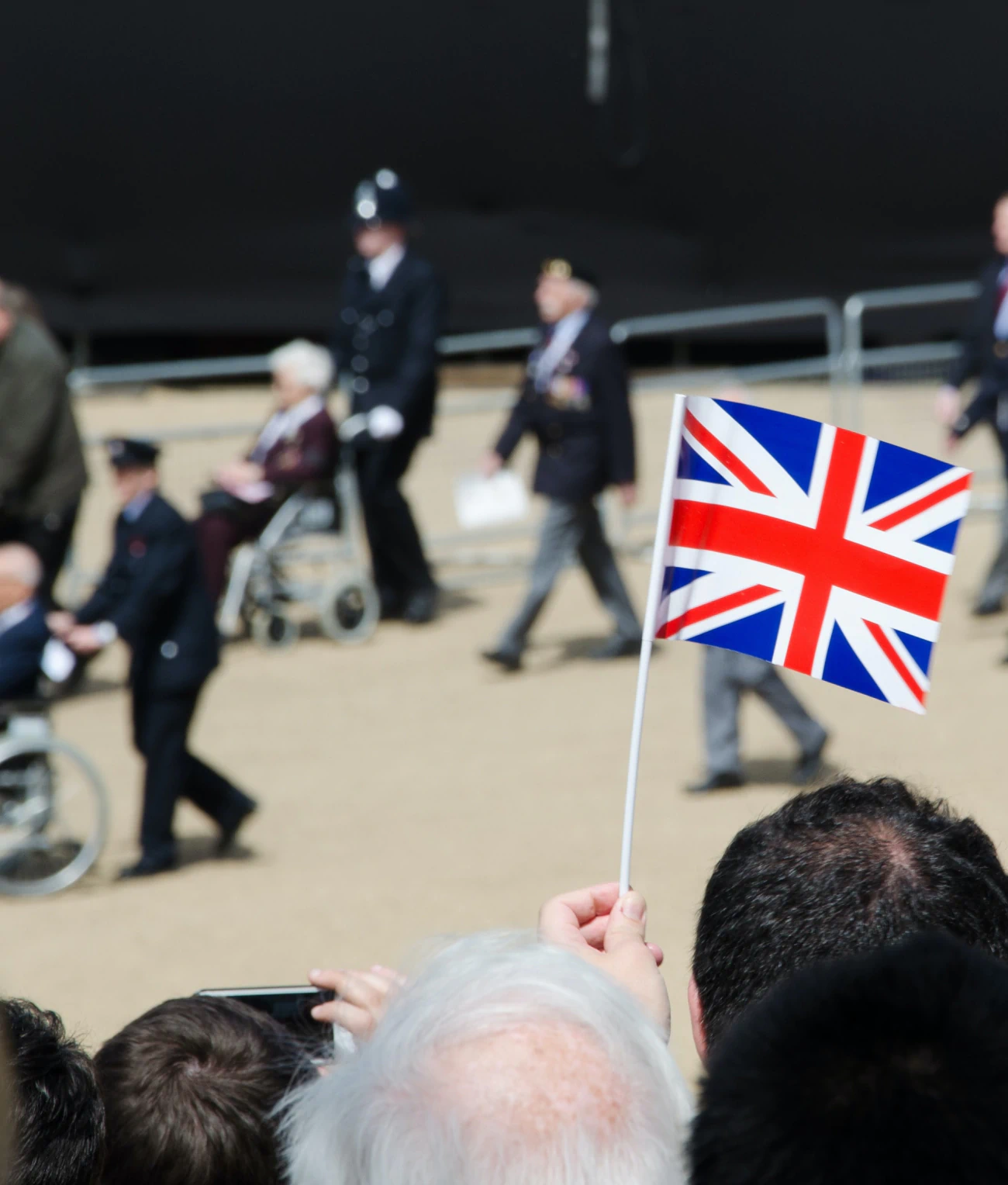 A small Union Jack flag held by a spectator in the foreground, with veterans and uniformed participants taking part in a remembrance procession in the background.