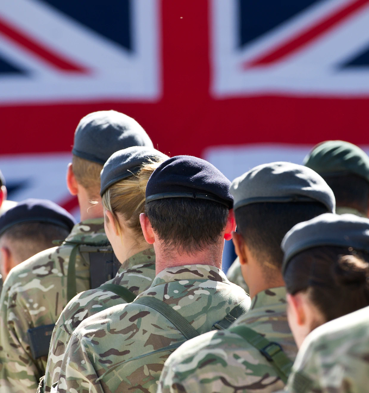 Uniformed British military personnel standing in formation, seen from behind, with a large Union Jack flag in the background.